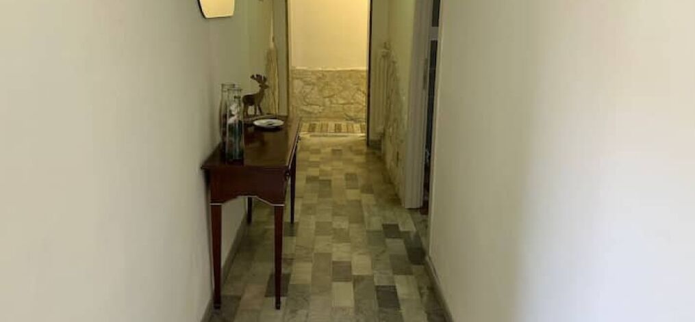 Un estrecho pasillo con piso a cuadros, una mesa de madera con un florero y un plato, y una pared de piedra al fondo.