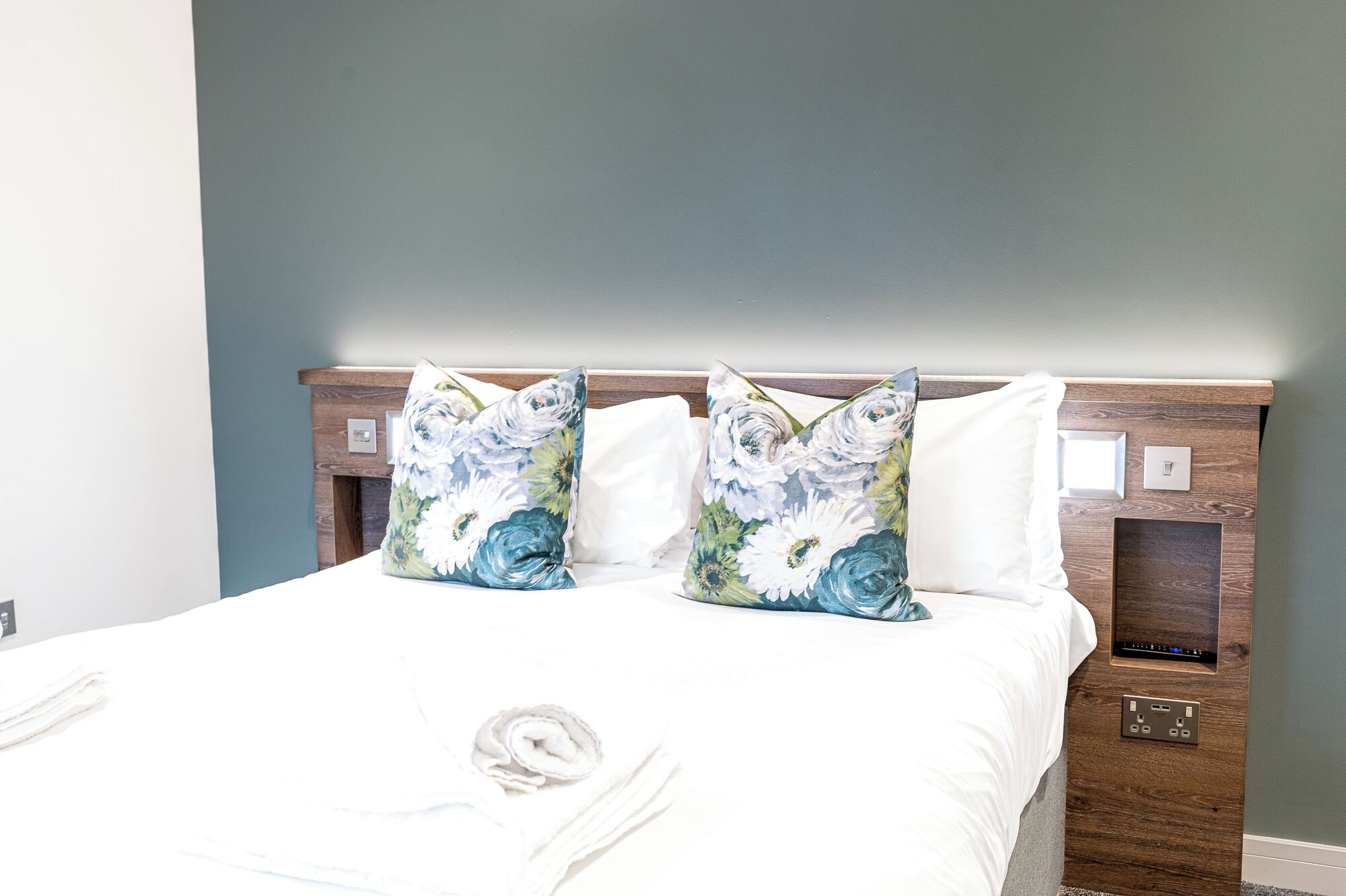 A neatly made bed with floral pillows, a wooden headboard, and a bedside table with a power outlet.