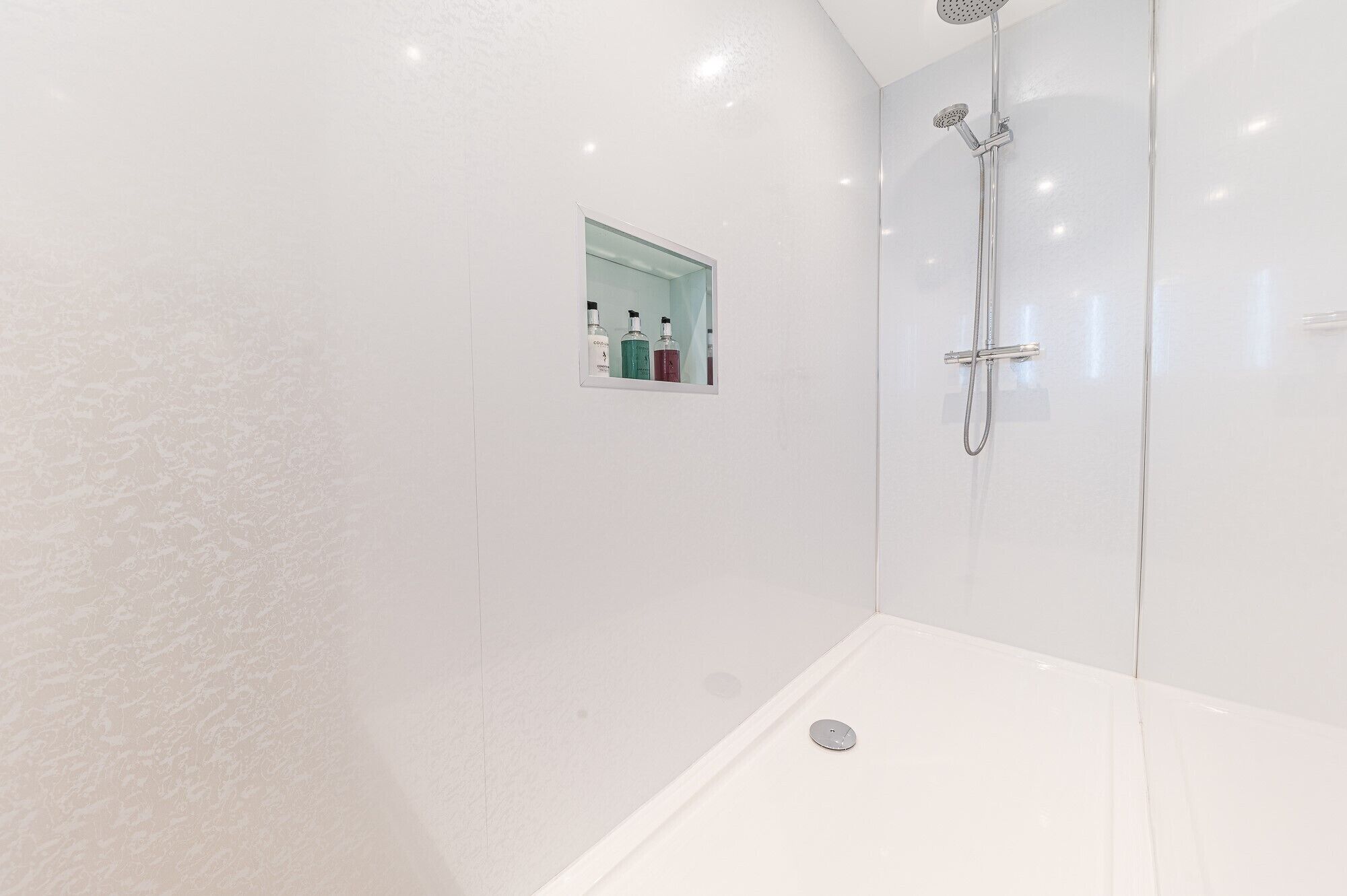 Ein modernes Badezimmer mit Duschkabine aus Glas, einer kleinen Ablage mit Pflegeprodukten und einem weißen Boden.
