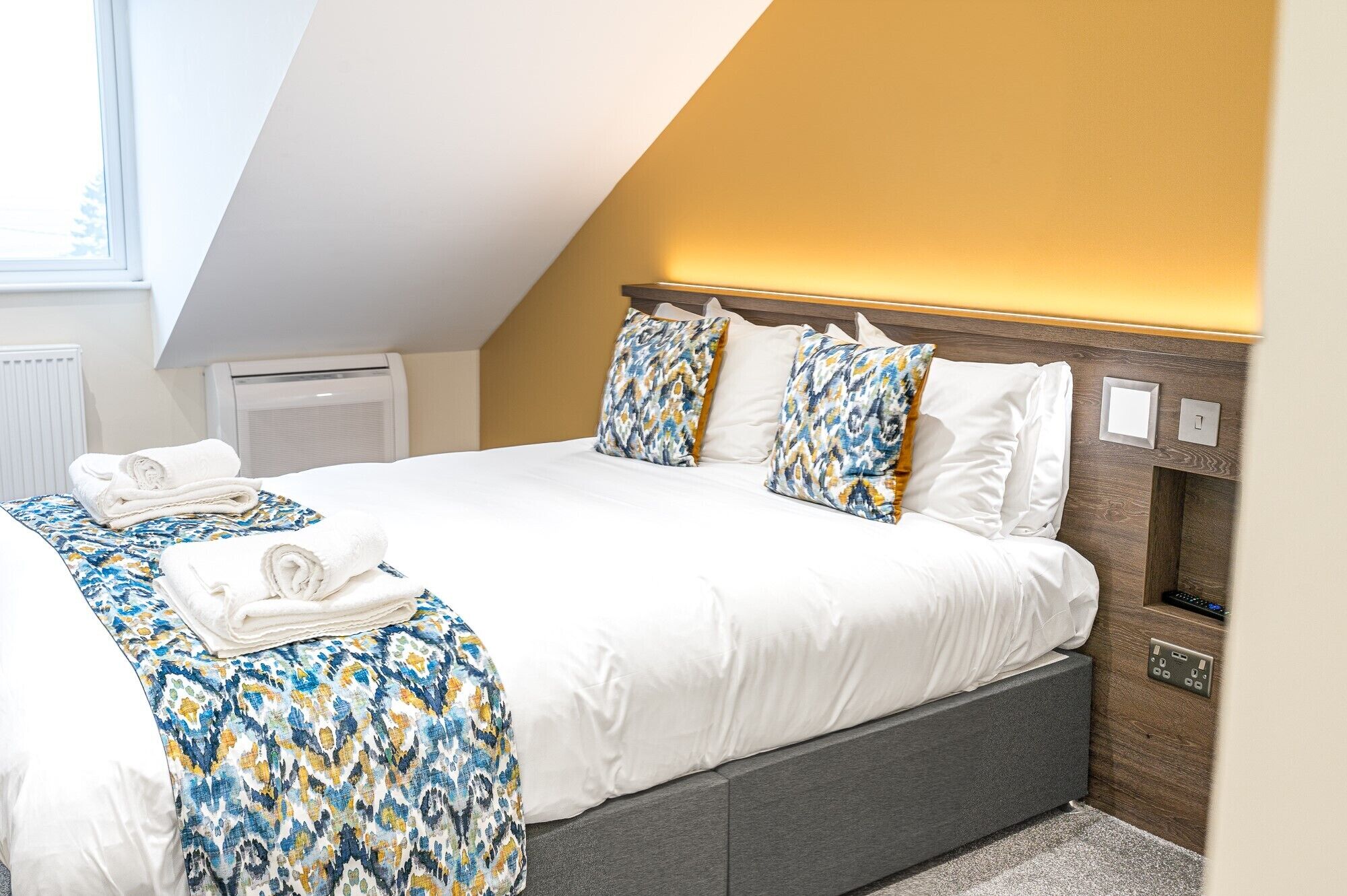 A neatly made bed with white linens and patterned pillows, a bedside table with a lamp, and a window with blinds.