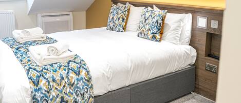 A neatly made bed with white linens and patterned pillows, a bedside table with a lamp, and a window with blinds.