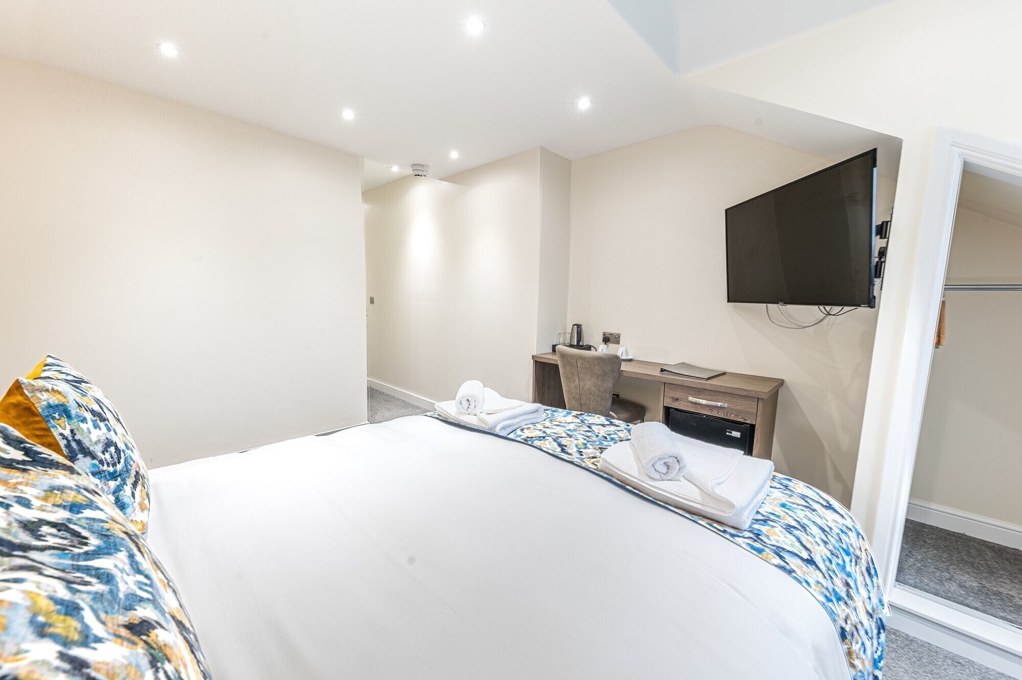 A hotel room with a bed, a desk, a chair, a flat-screen TV mounted on the wall, and a small kitchenette area.
