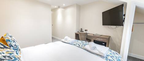 A hotel room with a bed, a desk, a chair, a flat-screen TV mounted on the wall, and a small kitchenette area.