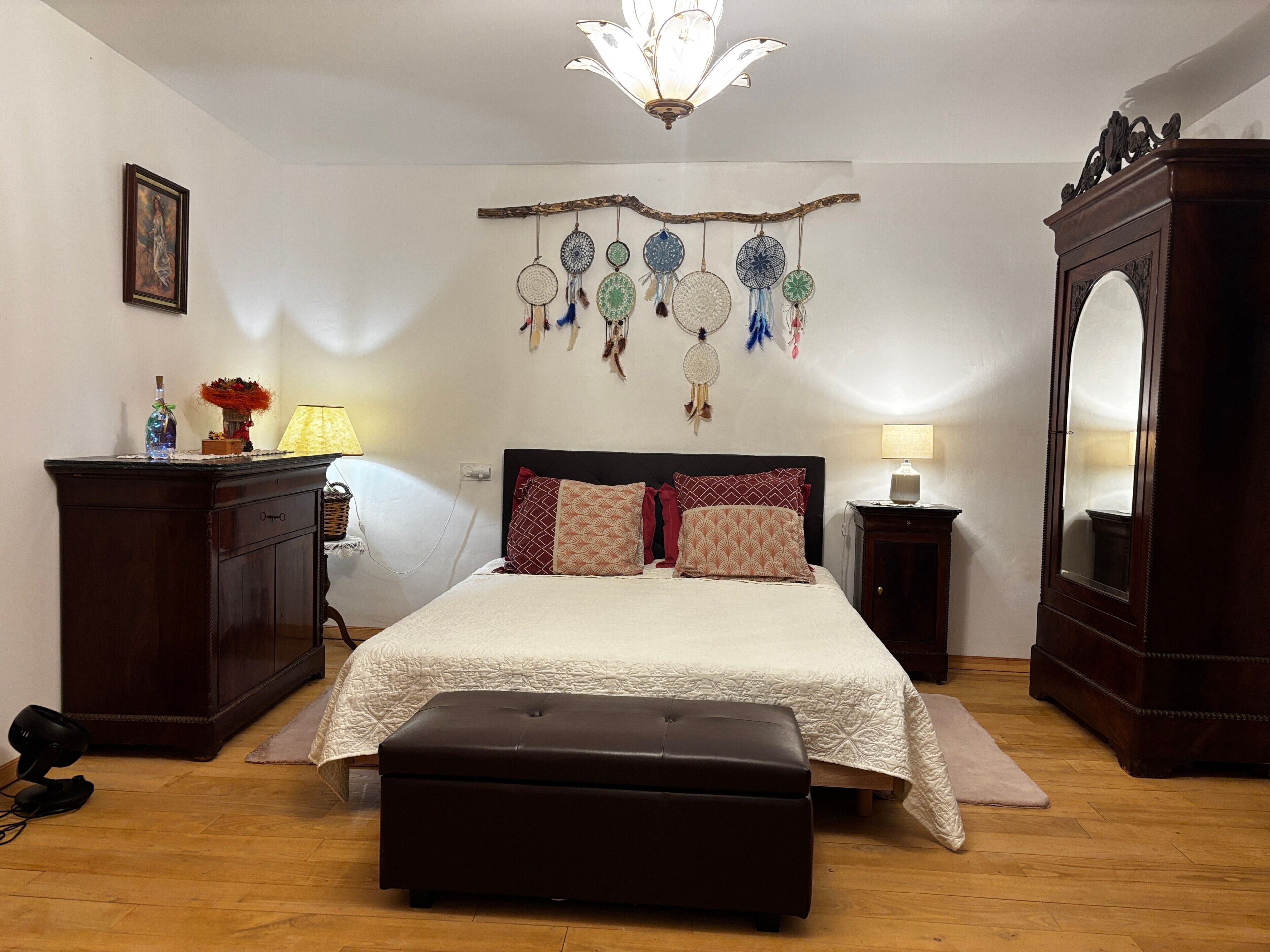 Quarto com cama, mesas de cabeceira, cômoda, espelho, lustre e decorações nas paredes.