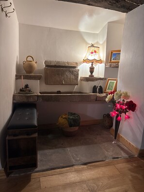 Een rustieke keuken met een stenen spoelbak, een houten bankje, een hangende pot, een kaars, een lamp en een vaas met bloemen.