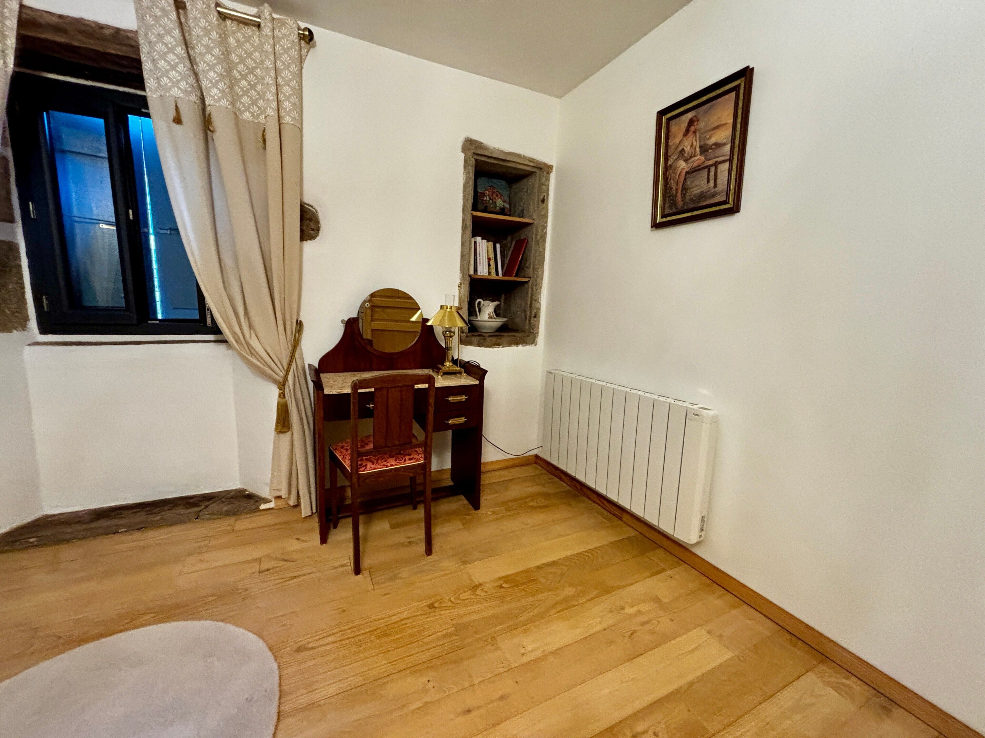 A room with a wooden desk, a chair, a radiator, a window with curtains, and a shelf with books.