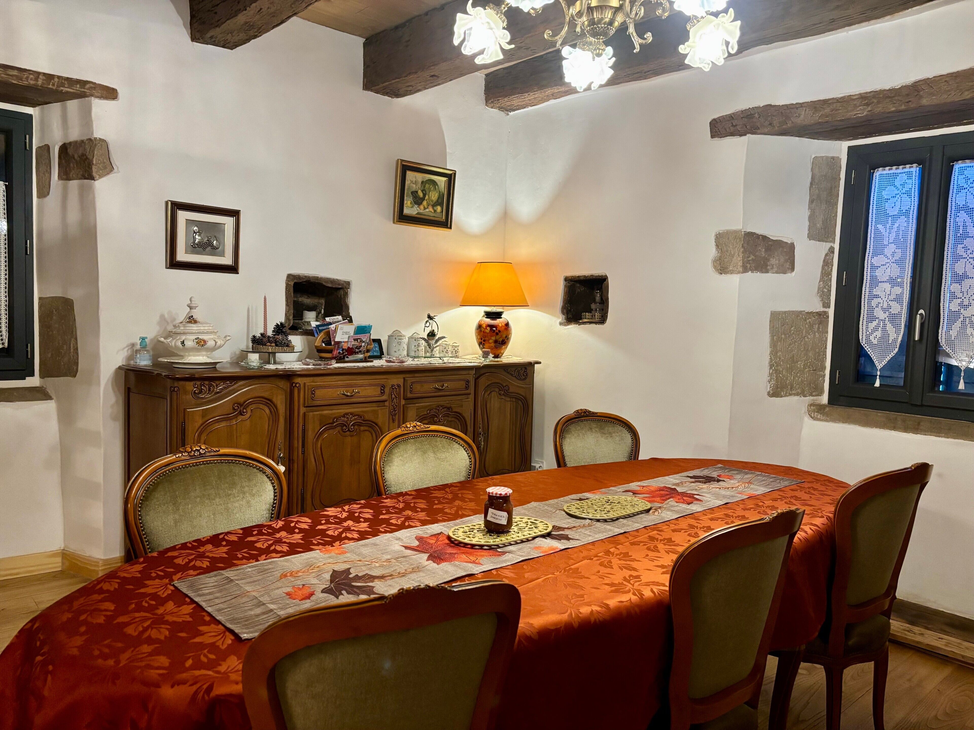 A dining area with a wooden table, chairs, a chandelier, and a window with lace curtains.