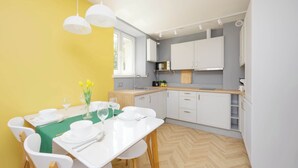 Una cocina moderna con armarios blancos, piso de madera en patrón de espiga y una zona de comedor para cuatro personas con sillas blancas y mantel verde.