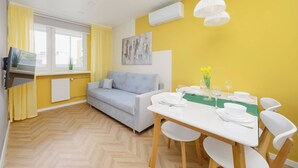 Un moderno salón con sofá, mesa de comedor y sillas. Hay un televisor sobre un mueble bajo, un cuadro en la pared y una ventana con cortinas.