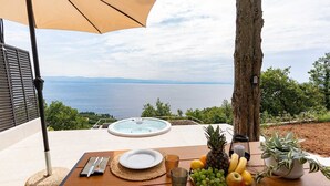 A table set outdoors with a hot tub, a scenic view of the sea, and a fruit basket.