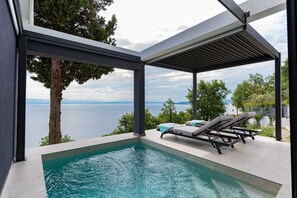 A modern outdoor pool area with a view of the sea, lounge chairs, and a pergola structure.
