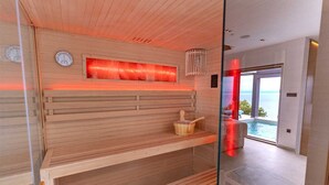 A sauna with wooden benches and a red-lit panel, a wooden bucket, and a wall-mounted thermometer.