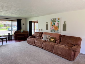 A living room with a brown leather sofa, a piano, a table, and a large window with a view of the street. - The Pelikan Retreat, Unique & Spacious Home, Large Yard (Missoula)