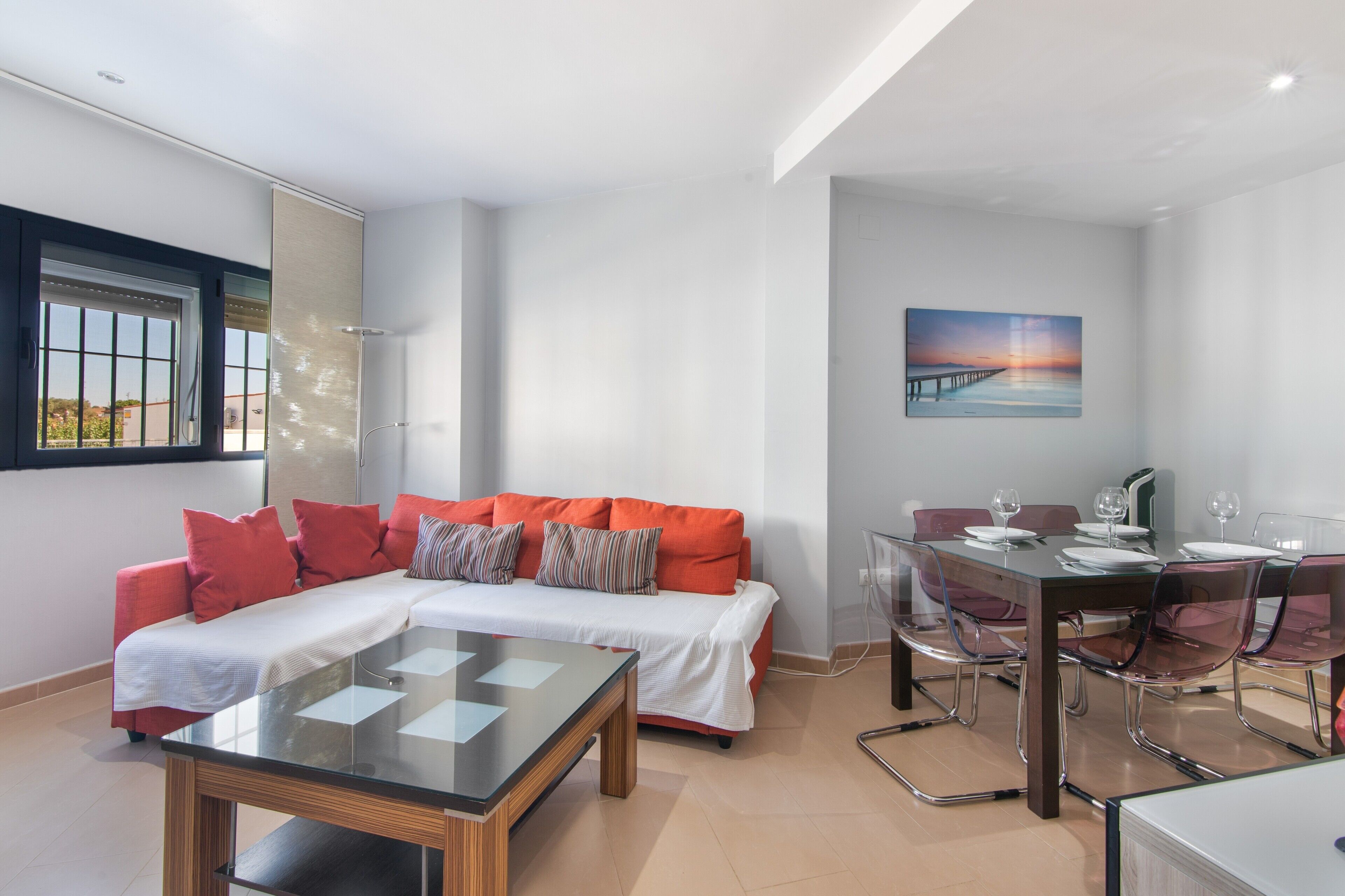 Uma sala de estar moderna com uma mesa de jantar de vidro, um sofá vermelho com almofadas brancas e um quadro na parede.