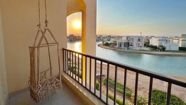 A balcony with a hanging chair, a view of a river, and modern buildings in the background. - Xuru Stays I Eclectic 2 BDR Twin House @ Tawila El Gouna (El Gouna)