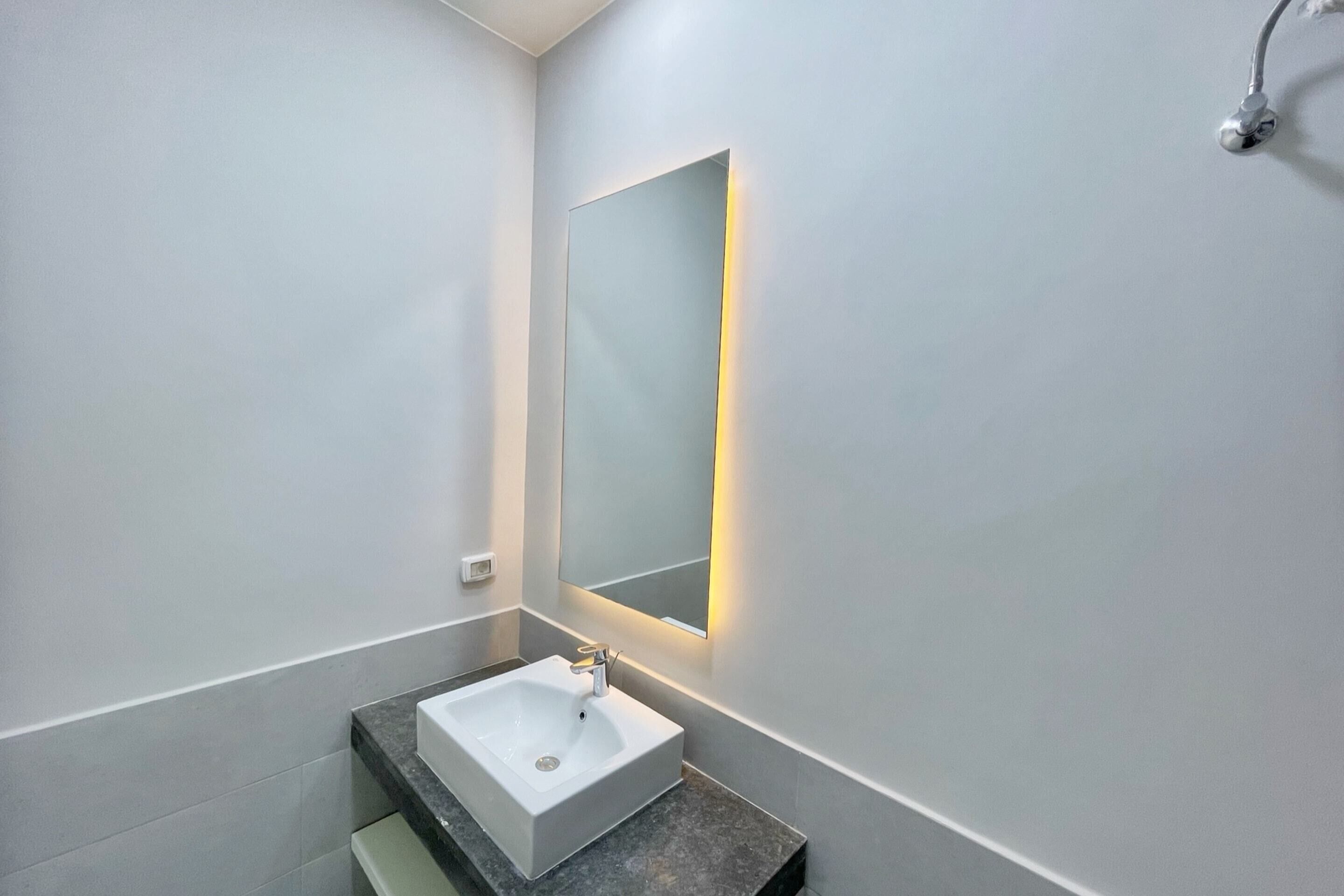 A modern bathroom with a white sink, a rectangular mirror with a wall-mounted light, and a showerhead.