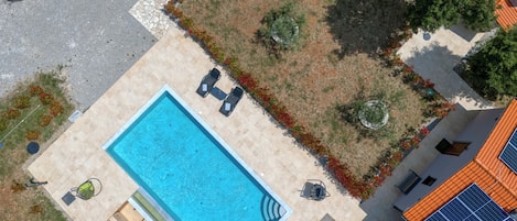 An aerial view of a swimming pool area with a tiled deck, lounge chairs, and a small garden with red flowers.