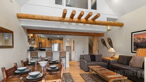 A spacious loft with a high ceiling, exposed wooden beams, a dining area with a wooden table and chairs, a gray sofa, and a kitchen in the background.