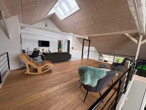 A spacious loft with a high ceiling, wooden flooring, a grey sofa, a wooden rocking chair, and a view of the outdoors through a skylight. - 4 Star Holiday Home in Henne (Henne)