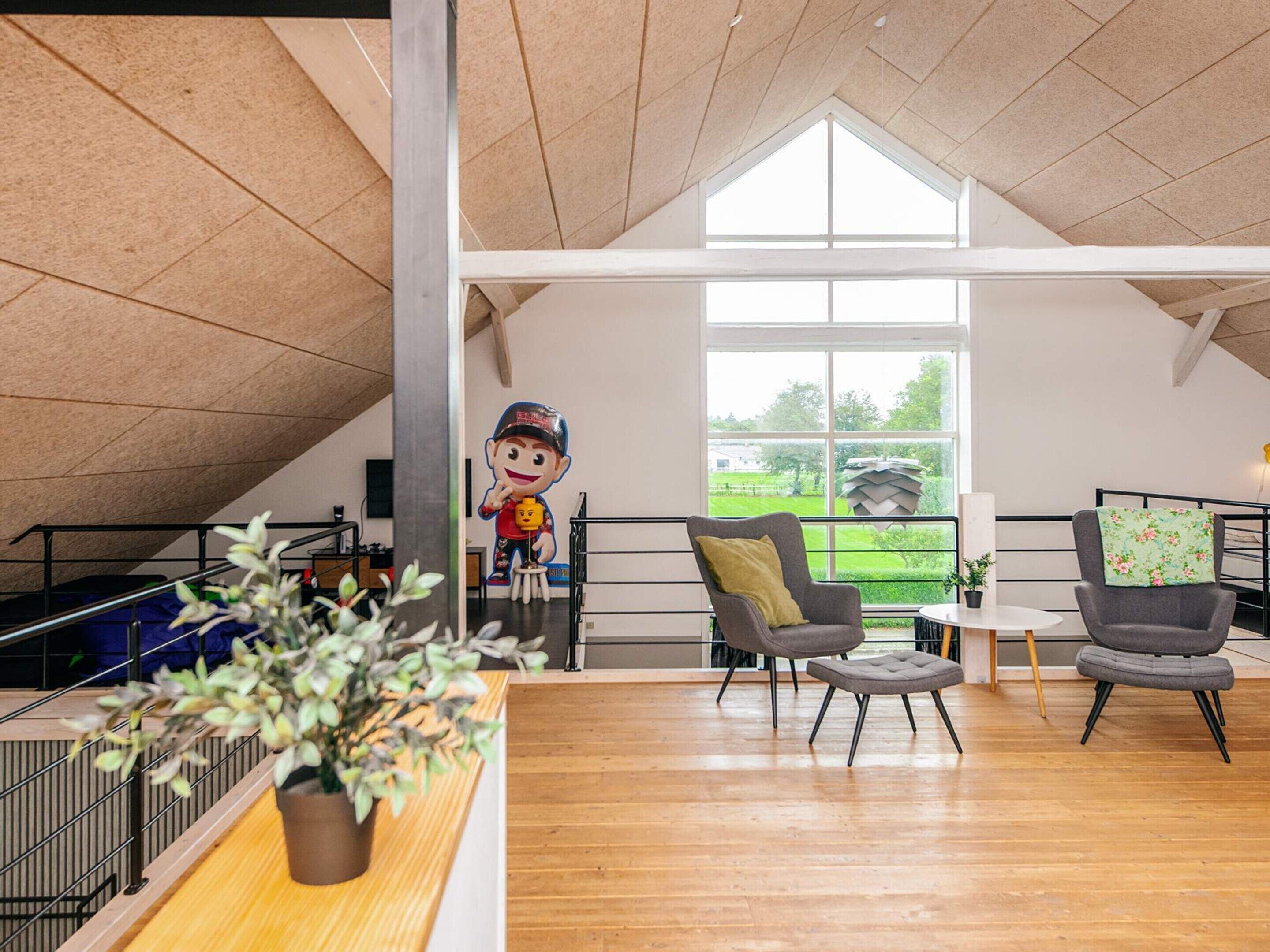 A spacious loft-style room with a high ceiling, large windows, and a view of a green landscape. There are two grey armchairs and a small table with a plant.