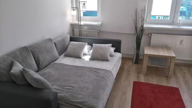 A modern bedroom with a grey sofa, a bed with grey bedding, a red rug, and a view of the sea through the window.