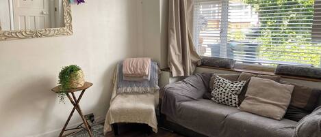 A cozy living room with a sofa, a small table with a plant, a decorative mirror, and a window with blinds.