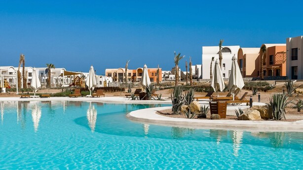 A resort pool area with clear blue water, palm trees, and modern buildings in the background. - Xuru Stays I Cool & Curated 1bdr @ Kamaran El Gouna (El Gouna)