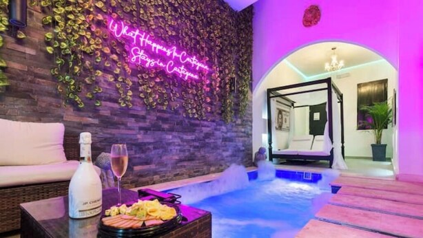A spa area with a hot tub, a neon sign, a bottle of champagne, and a tray of snacks.