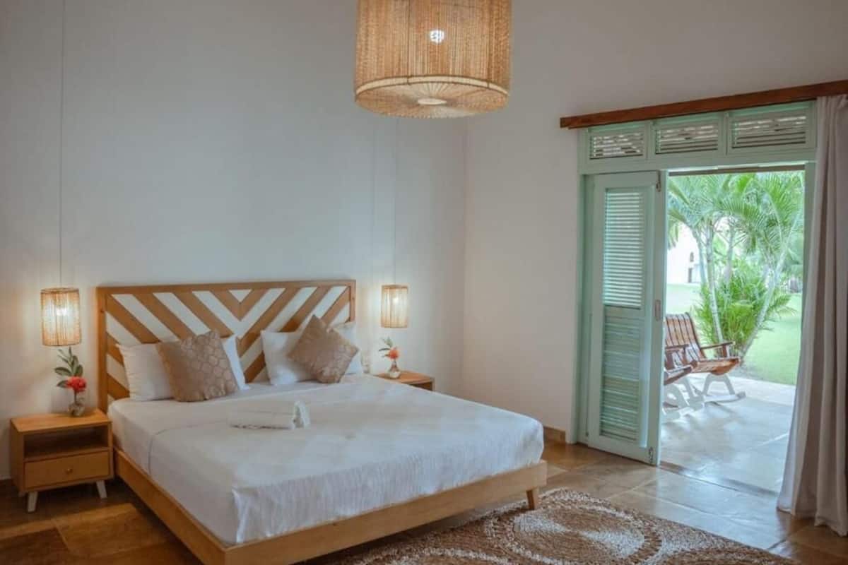 A bedroom with a wooden bed, two bedside lamps, a woven ceiling light, and a view of a patio with a chair and plants.