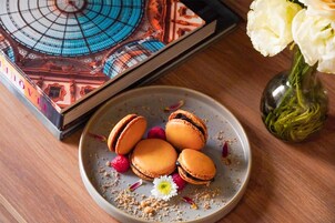 Un plato de macarons con una flor blanca y migas esparcidas, un florero con flores blancas y un libro con cubierta azul sobre una mesa de madera.