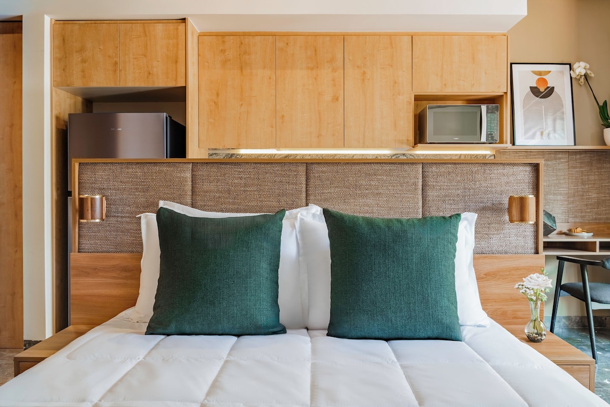 A hotel room with a bed, a kitchenette, a television, and a small table with a vase.