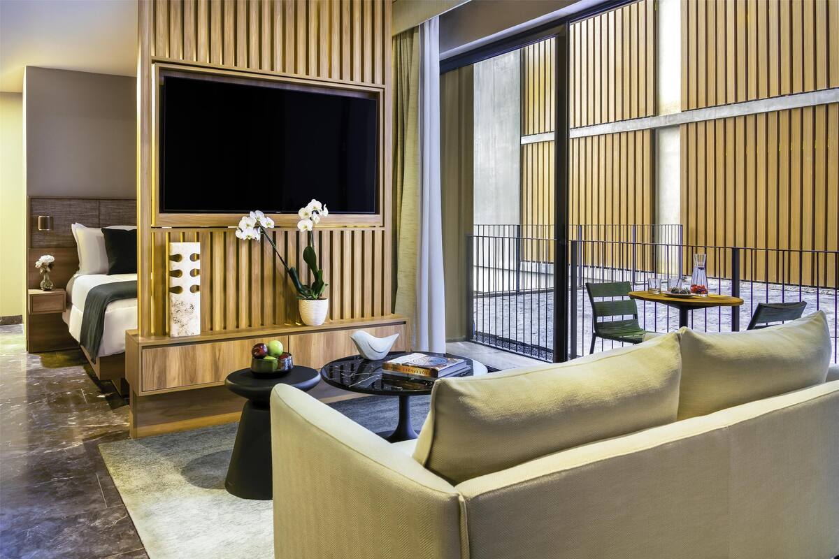 Moderni hotellihuone, jossa on suuri televisio, sohva, pieni pöytä ja parveke, jossa on tuolit.