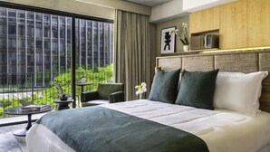 Habitación de hotel moderna con una cama grande, vistas a zonas verdes a través de ventanales de gran altura, una mesita con un libro y una planta en maceta.