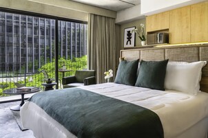 A modern hotel room with a large bed, a view of greenery through floor-to-ceiling windows, a small table with a book, and a potted plant.