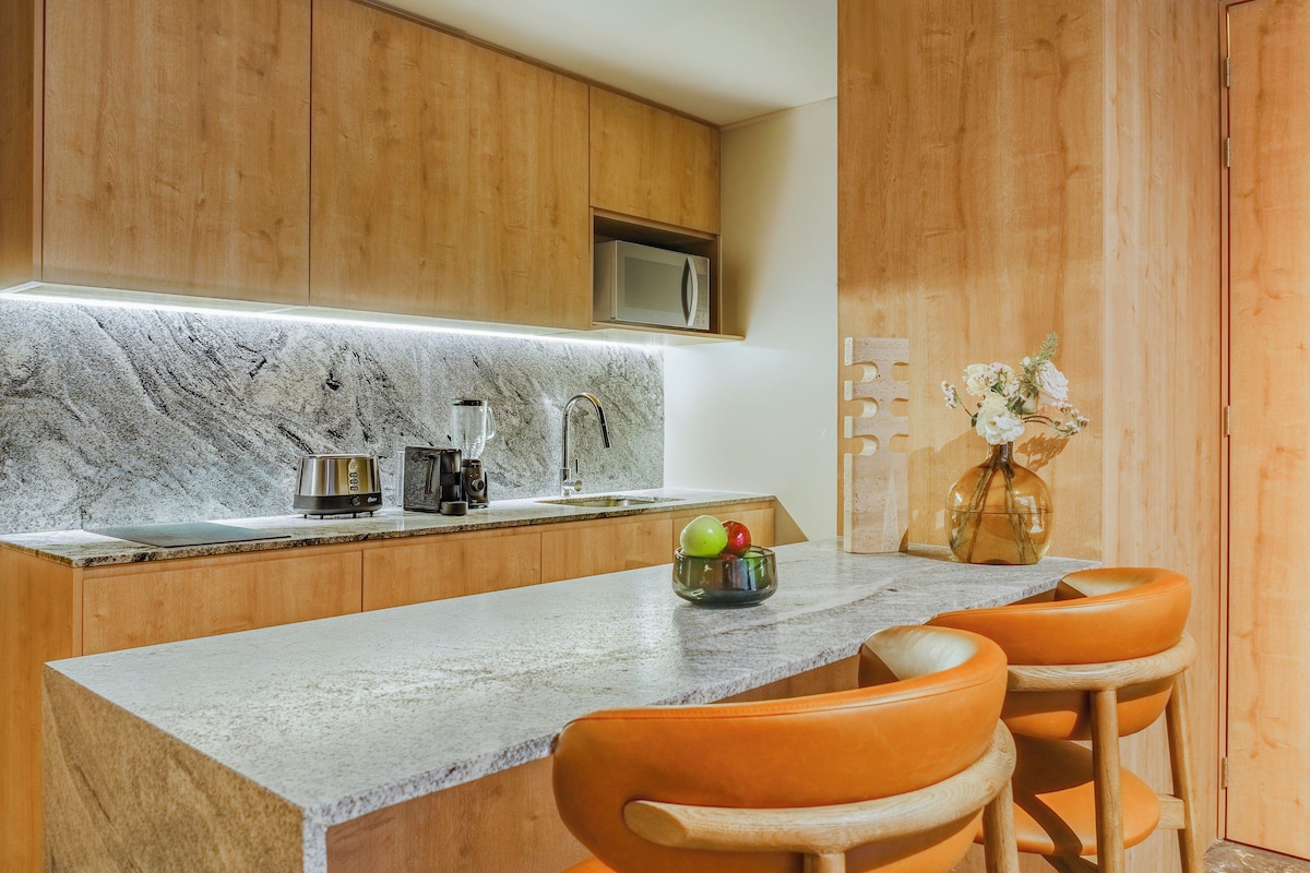 A kitchen with wooden cabinets, a marble countertop, a microwave, a sink, and two orange stools.