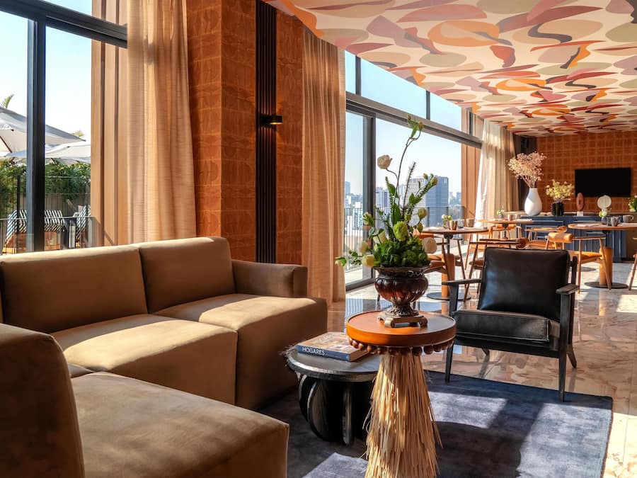 Un moderno hall de hotel con un sofá grande, una mesa redonda con un florero y vistas a la ciudad a través de amplias ventanas.