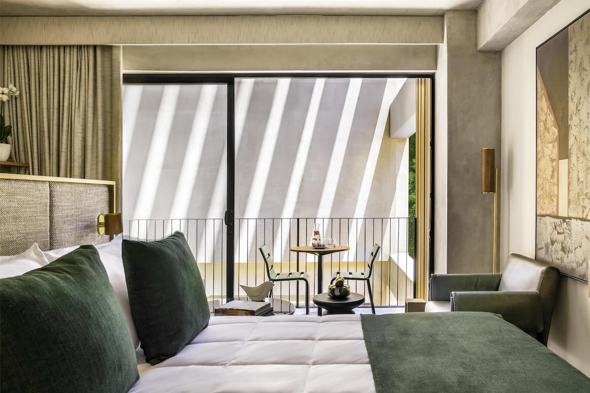 Moderni hotellihuone, jossa on suuri sänky, vihreät tyynyt, pieni pöytä tuoleineen sekä parveke, jossa on raidalliset verhot.