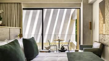 Habitación de hotel moderna con una cama grande, almohadillas verdes, una mesita con sillas y un balcón con cortinas a rayas.