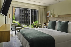 A hotel room with a large bed, a desk, a chair, a TV, and a balcony with a view of greenery.