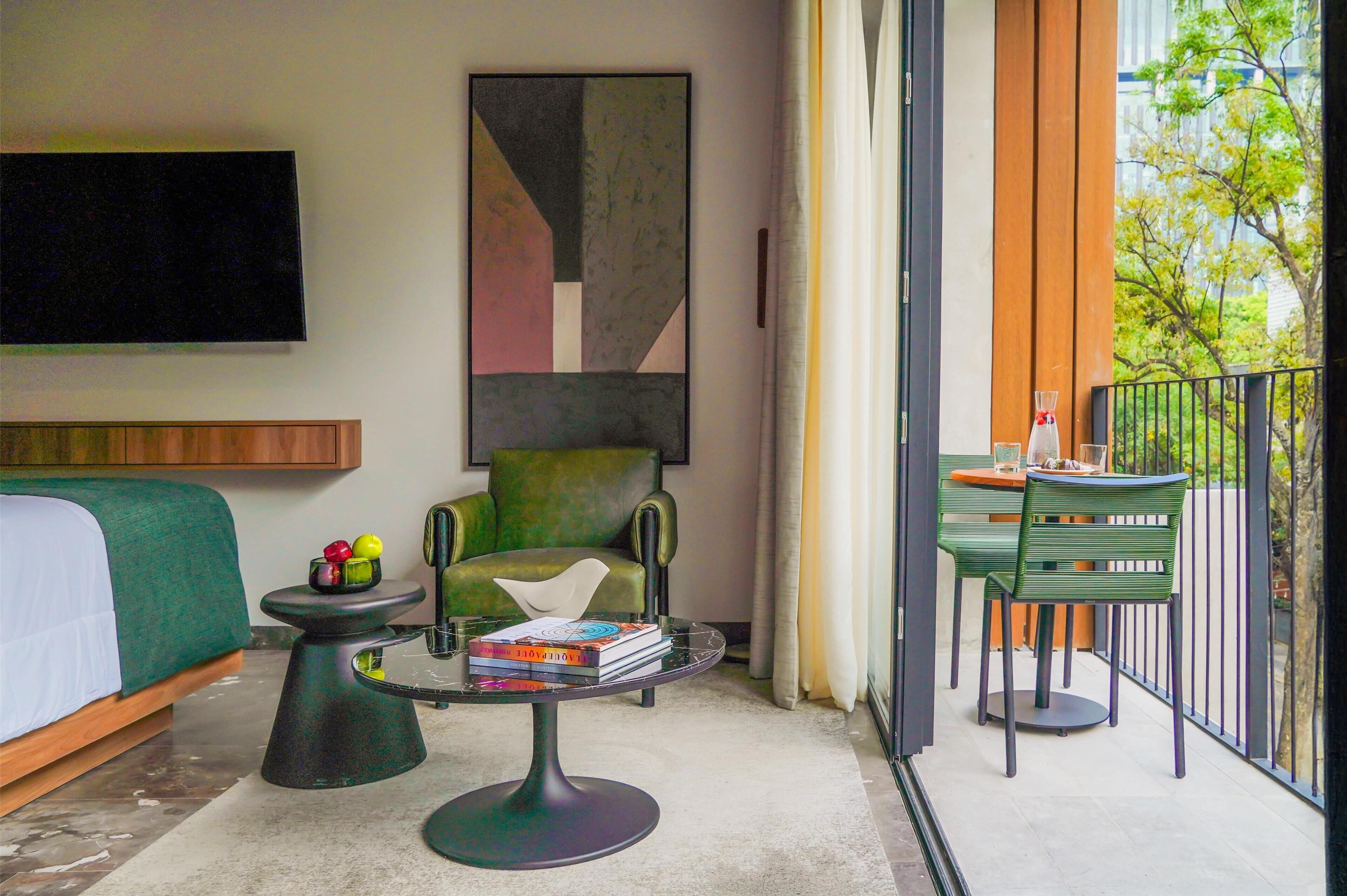 Une chambre d’hôtel moderne avec un lit, un fauteuil vert, une petite table avec des livres et un balcon équipé d’une table et de chaises.