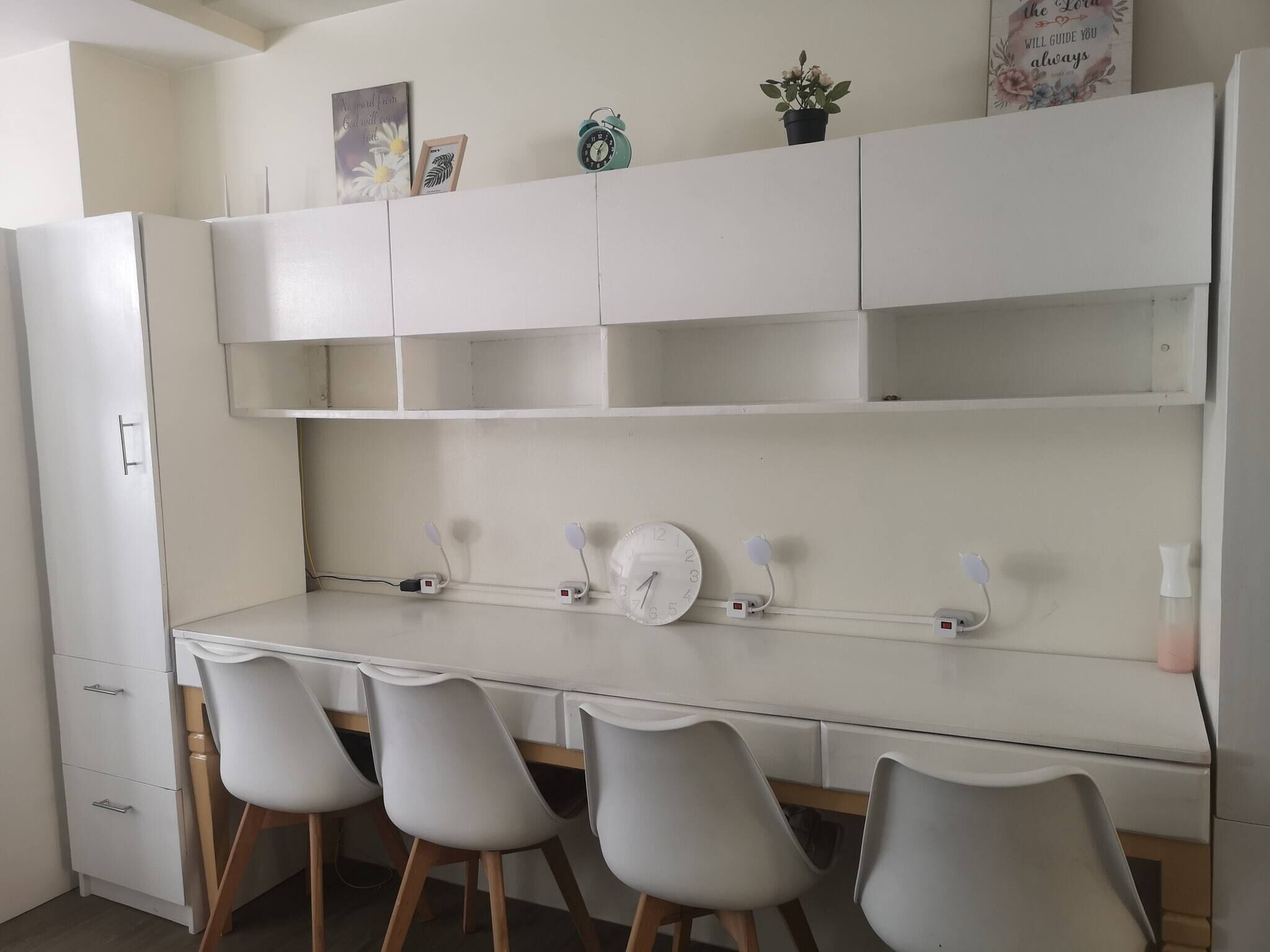 A kitchen with white cabinets, a white countertop, and four grey chairs. There is a clock and a small potted plant on the countertop.