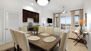 A dining area with a wooden table, beige chairs, and a chandelier. There is a kitchen in the background with dark cabinets and a flat-screen TV.