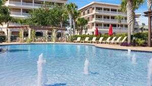 A swimming pool with water fountains, surrounded by lounge chairs and palm trees, with a hotel building in the background.