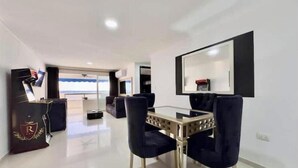 Una sala de estar moderna con una mesa de centro de vidrio, sillas azul oscuro, un televisor de pantalla plana montado en la pared y vistas al mar a través de amplios ventanales.