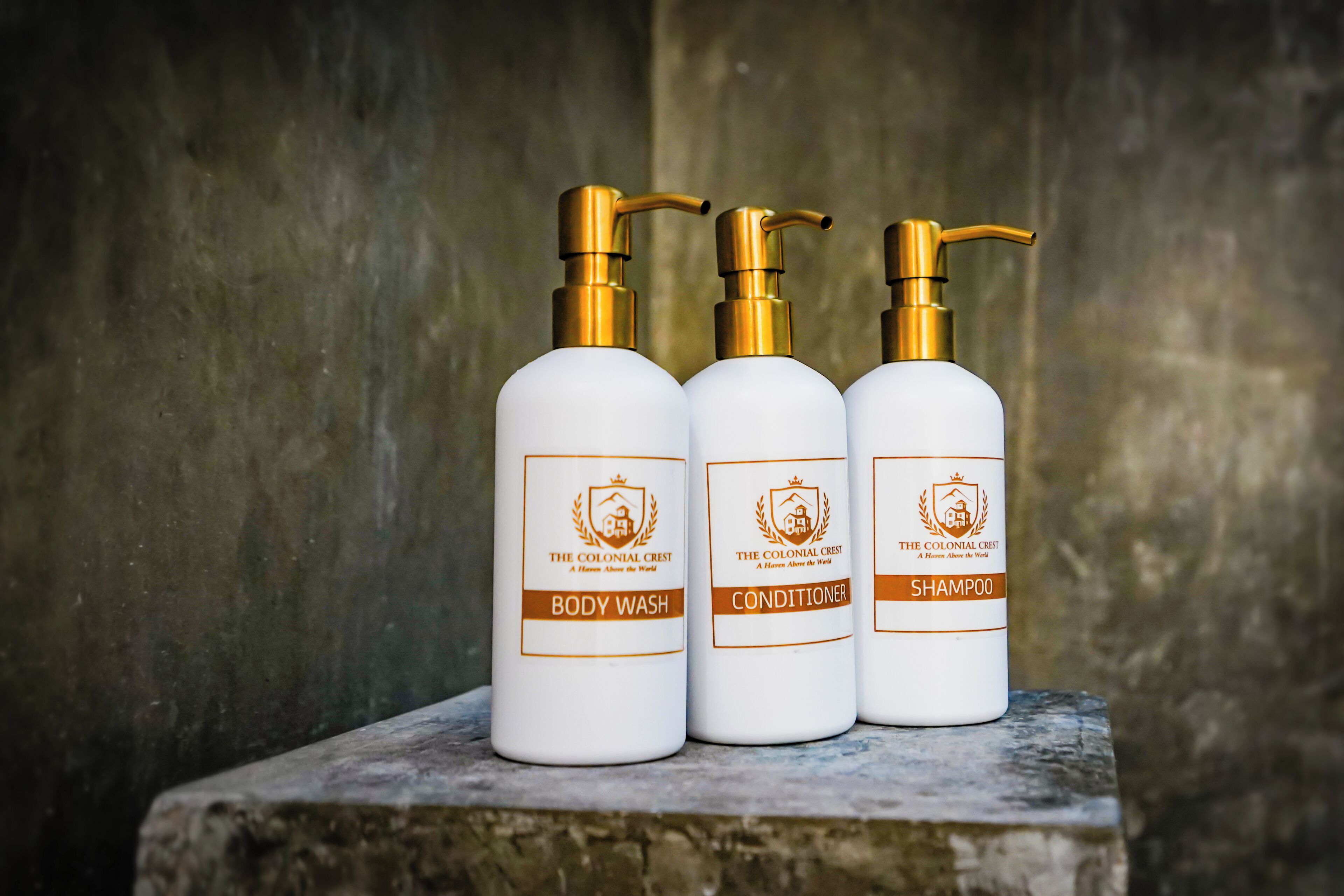Three bottles of The Colonial Crest body wash, conditioner, and shampoo on a stone ledge.