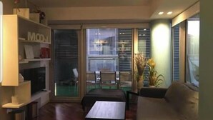 A modern living room with a sofa, a coffee table, and a bookshelf. There is a sliding glass door leading to an outdoor area with a table and chairs.