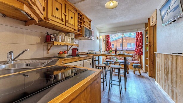 Een keuken met houten kasten, een roestvrijstalen spoelbak en een groot eiland. Er staan barkrukken en een tafel in de eetruimte, en er is een raam met gordijnen.