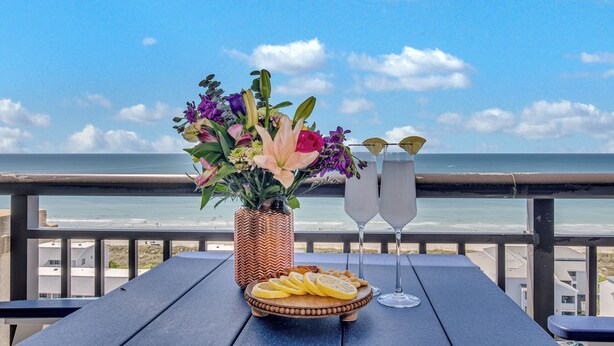 A table with a floral arrangement, a plate of lemon slices, and two glasses of a clear beverage, overlooking a beach. - 3BR- 14th floor- Oceanview Corner Condo (North Myrtle Beach)