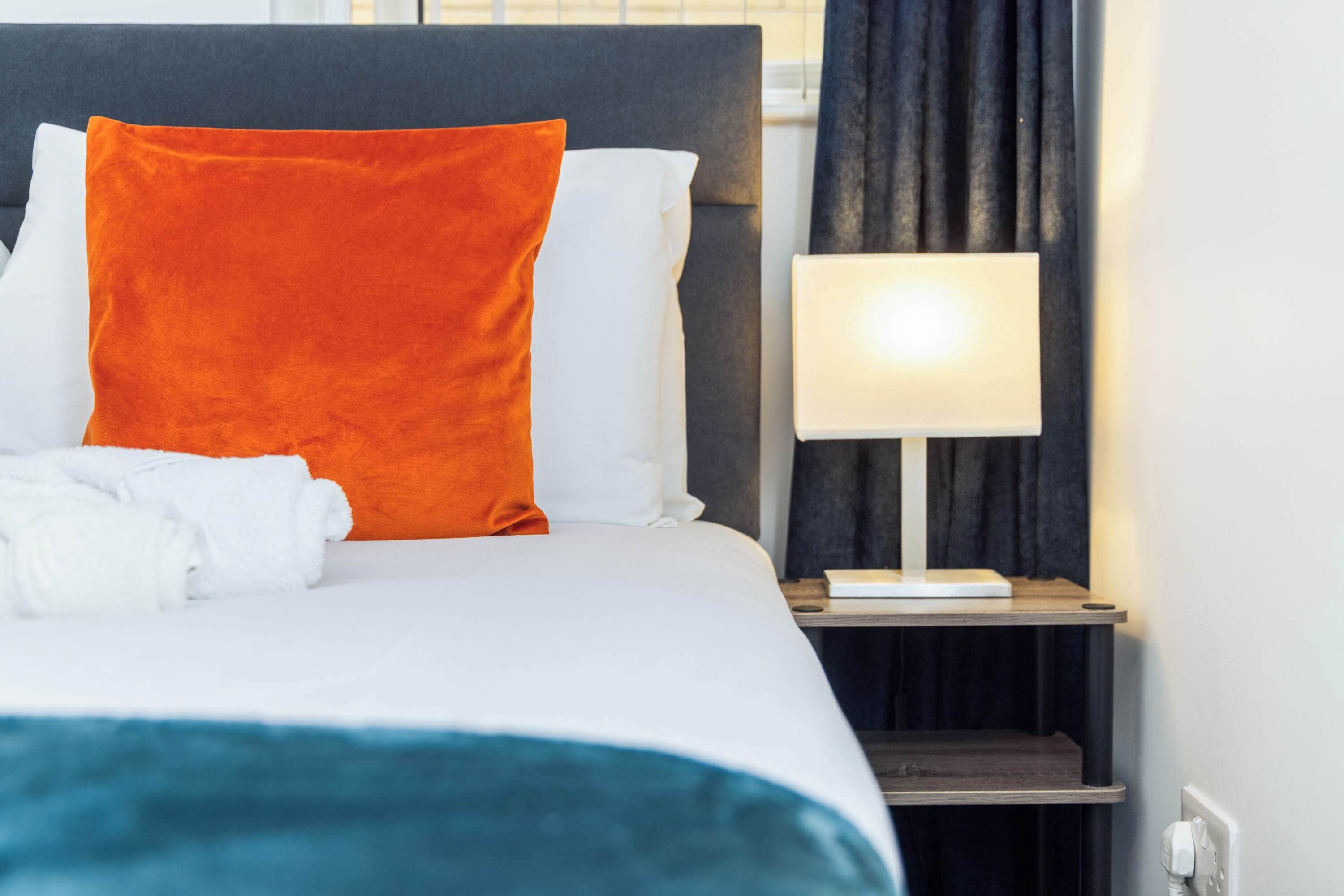 Un letto rifatto con cuscini bianchi e arancioni, una coperta turchese e un comodino con una lampada.