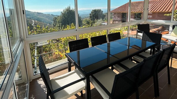 Une terrasse couverte avec une table et des chaises en verre, de grandes fenêtres offrant une vue sur la campagne.